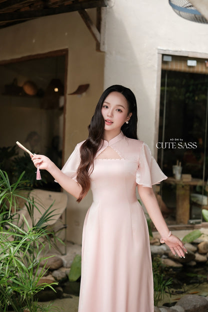 A woman wearing a pale pink traditional-style dress stands outdoors in front of a building with plants and a window. She is holding a small pink tassel accessory in one hand, and the text 'CUTE SASS' appears on the right side of the image.