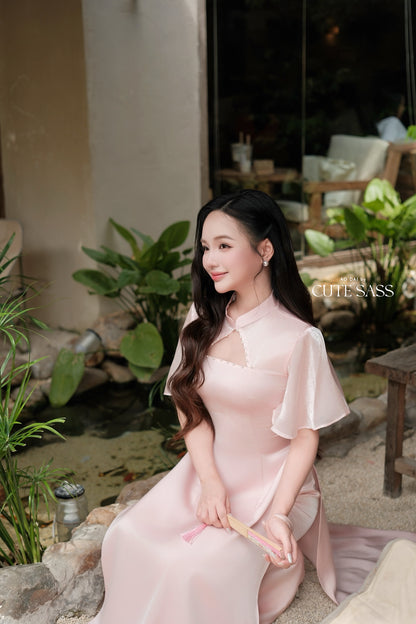 A woman wearing a light pink traditional Vietnamese ao dai dress sits in a garden courtyard setting. The dress features a high collar, short flutter sleeves, and a keyhole cutout at the chest.