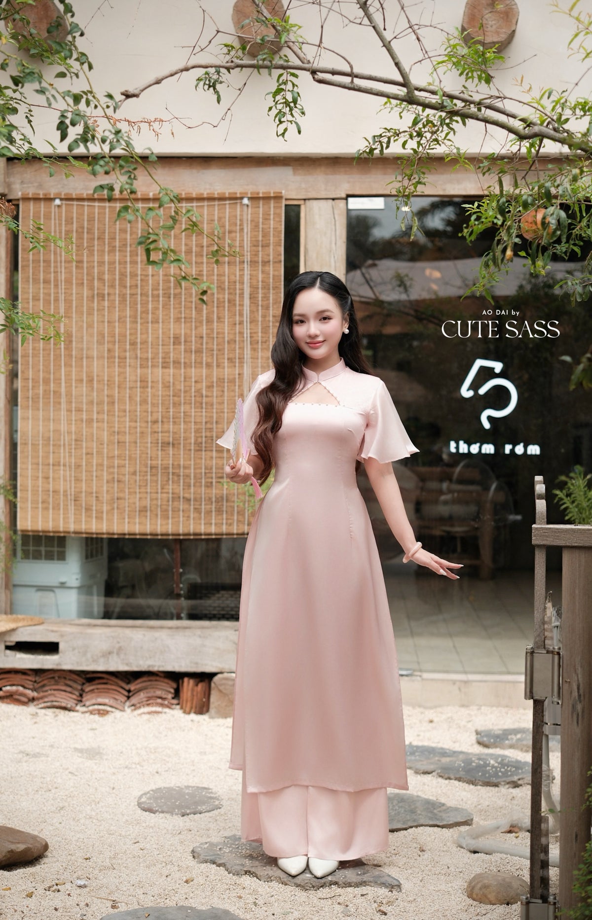 A woman wearing a light pink traditional Vietnamese ao dai dress stands outdoors in front of a cafe storefront with a bamboo screen and green foliage.