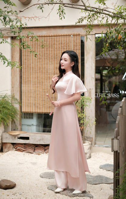 A woman wearing a light pink traditional Vietnamese ao dai dress stands in a courtyard setting with a bamboo screen and wooden door in the background.