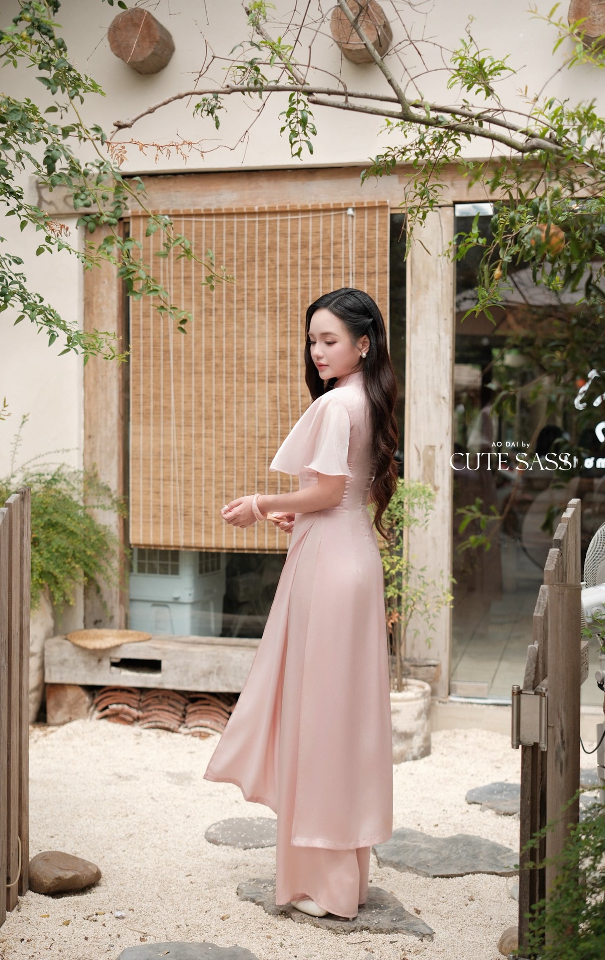 A woman wearing a light pink, short-sleeved maxi dress with a side cutout stands in a rustic courtyard setting. The background features a bamboo screen, wooden door, and stone path.